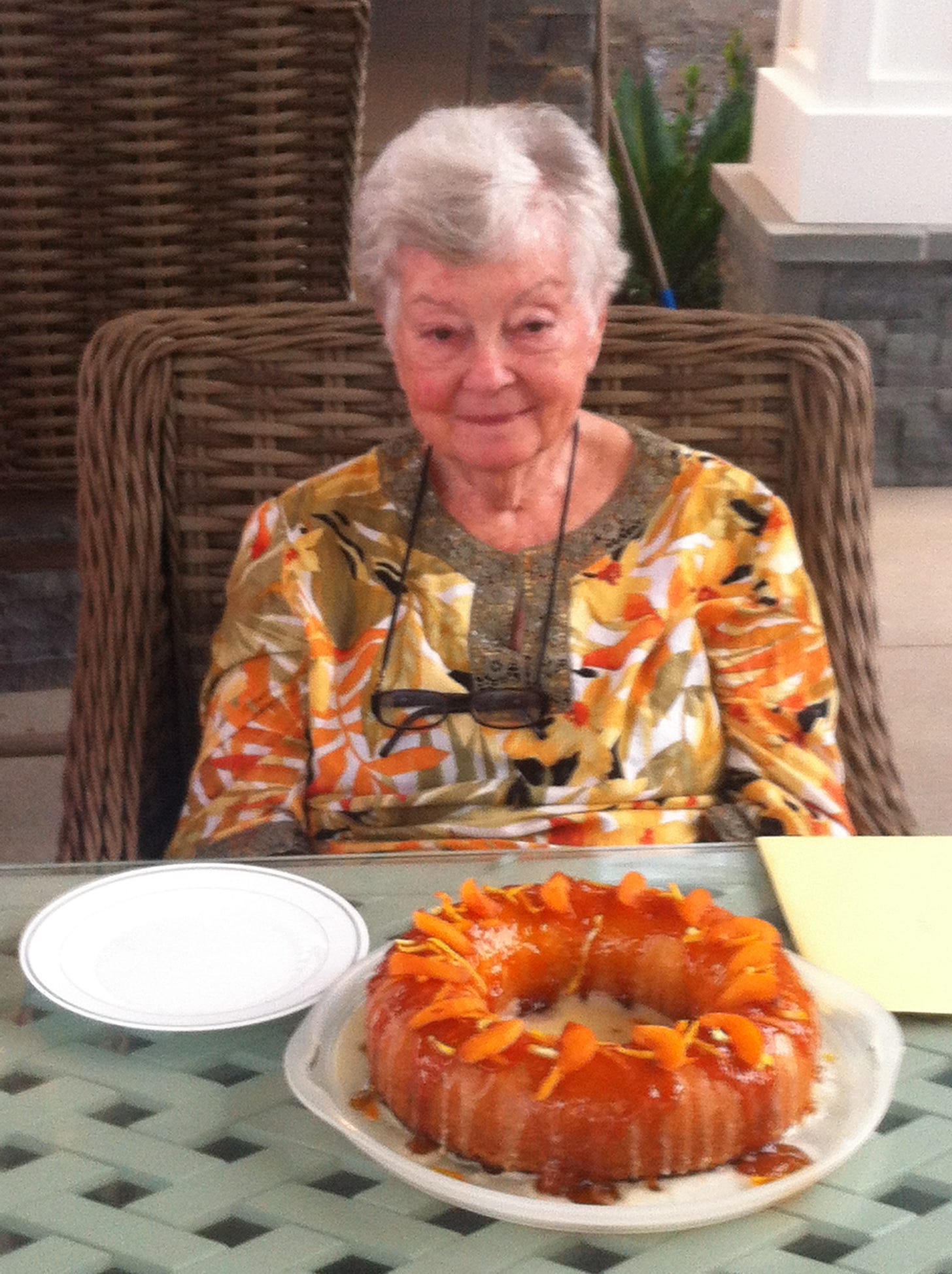 An older woman (84 years) sitting in a wicker chair with an apricot ring cake on the table in front of her.