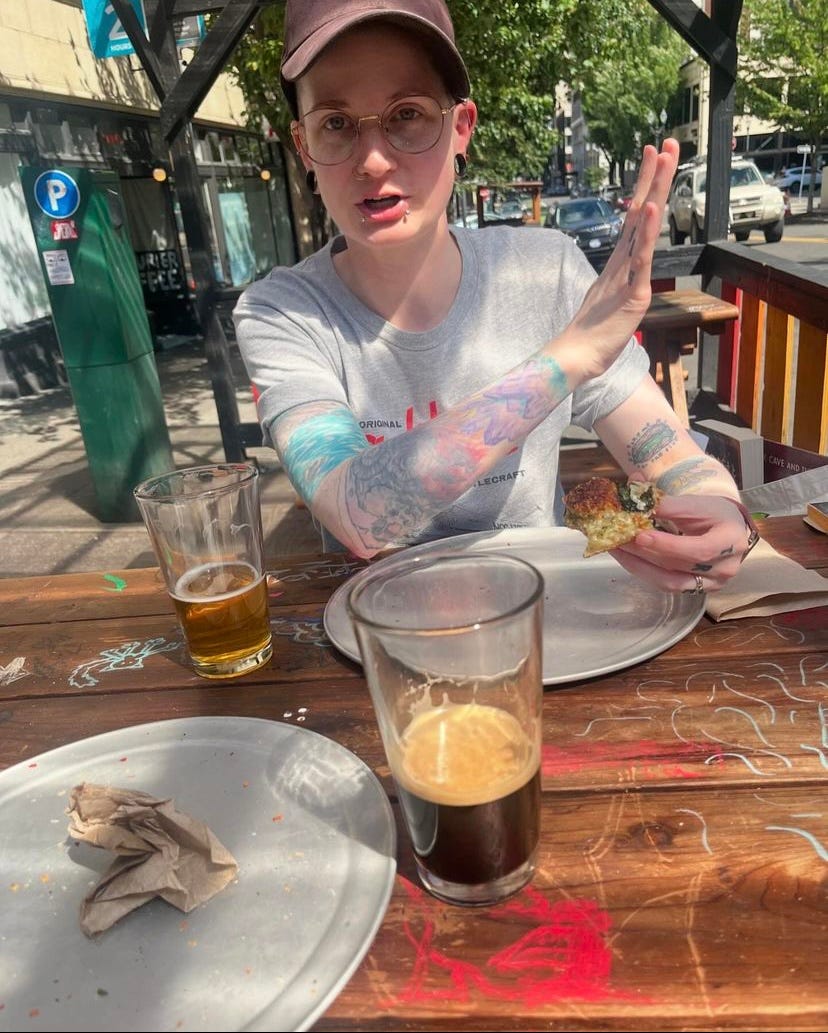 a photo of me, holding the remains of a piece of pizza in one hand and gesticulating with the other hand. I'm at an outdoor table, wood with drawings on it, metal pizza plates and nearly-empty glasses of beer on the table.