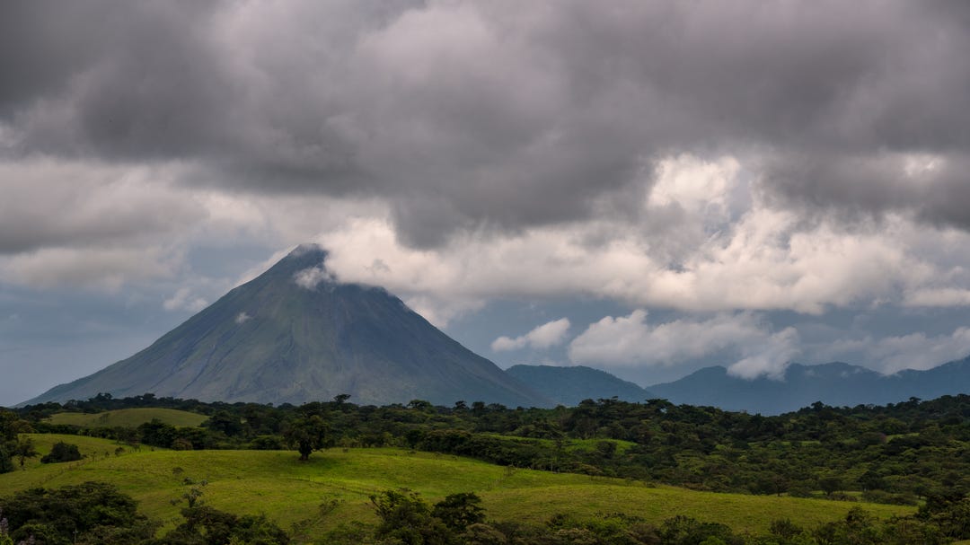 Arenal Volcano in Costa Rica with rolling green hills in front and cloudy sky above.