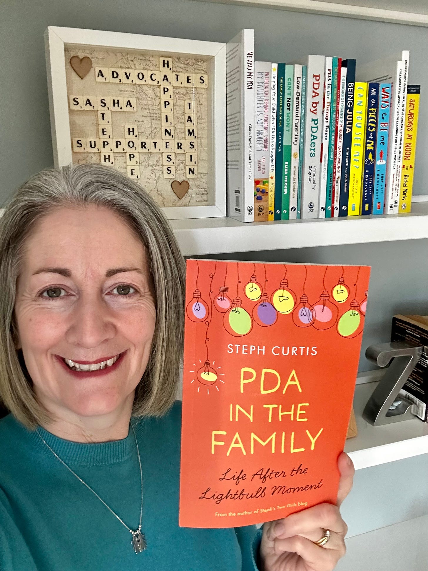 steph, middle aged woman with silver hair standing in front of shelves with pda books on holding up book with orange cover and title pda in the family