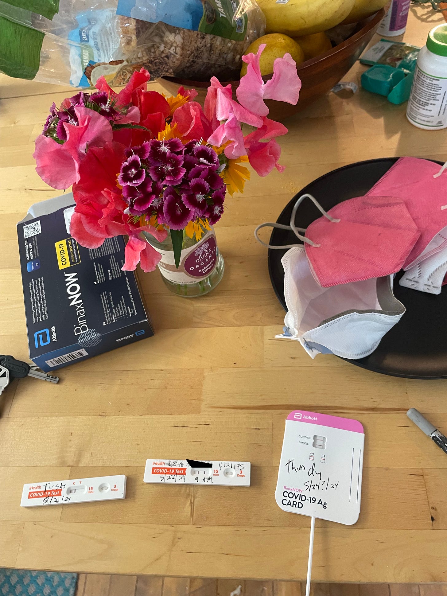 a wooden kitchen table holds a bouquet of wildflores in pinks, purples and yellow, next to bottles of supplements, a bowl holding pink KN-94 masks and a opened Binax NOW COVID 19 testing box. in front, there is a positive covid test with two lines and the date marked on it in black pen.