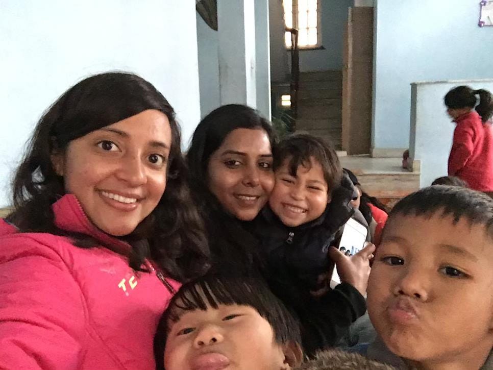 Two South Asian women in their 20s and 30s smile into the camera. Surrounding them are small Nepali children making various faces as they look at the camera. 