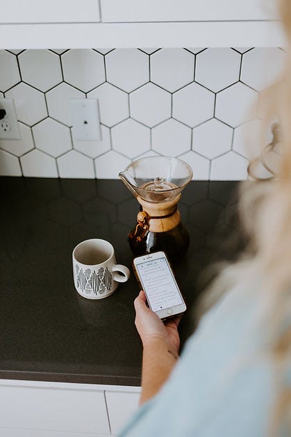 Image of an espresso maker, and coffee cup with a woman reading a message on her cellphone.
