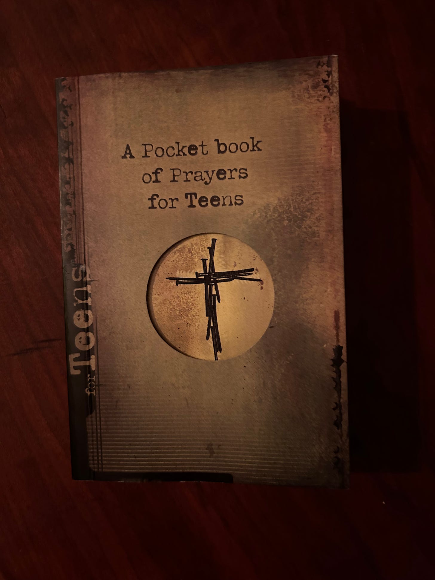 A little book entitled A Pocket Book of Prayers for Teens with an image of a cross made from nails. The book lies on a wooden table.