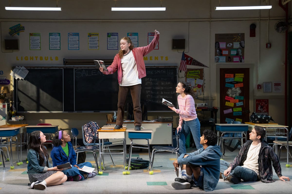 Sadie Sink as Shelby Holcomb in John Proctor is the Villain stands on a desk in a high school classroom reading from The Crucible as her classmates look on.
