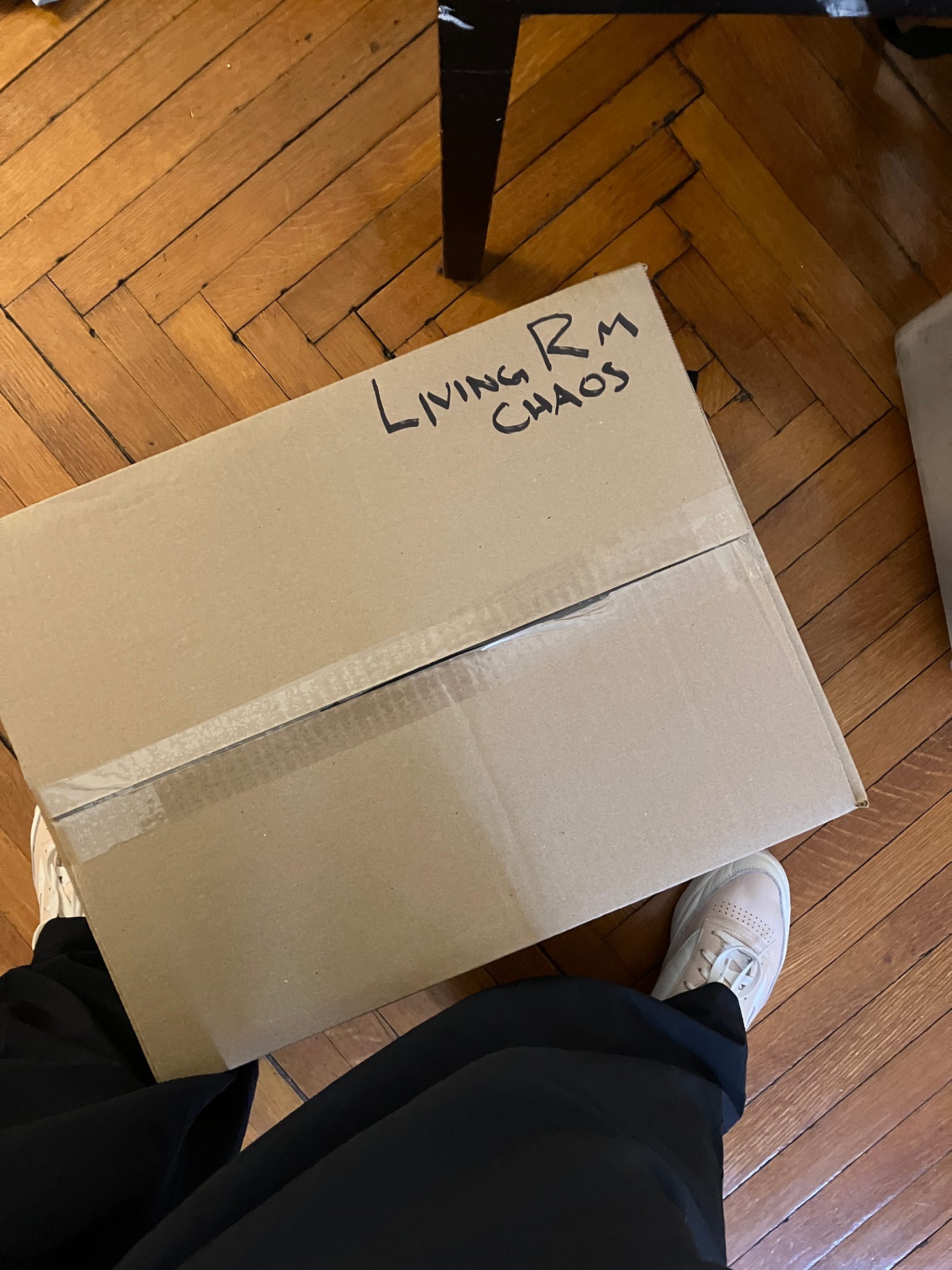 A sealed box on a hardwood floor labeled with Sharpie and all capital letters "LIVING RM CHAOS"