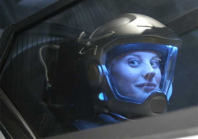 A white woman in a space helmet looking at the camera through the windshield of a spaceship. She looks kind of mischievous.