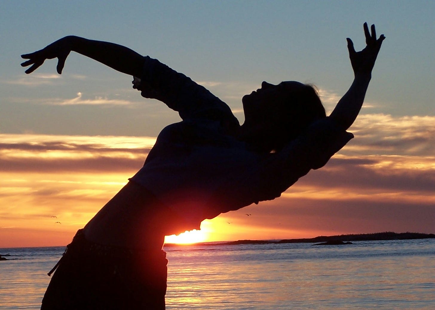 The author in a backbend with artistically placed dancer arms, in silhouette before the sun setting over the Pacific Ocean.