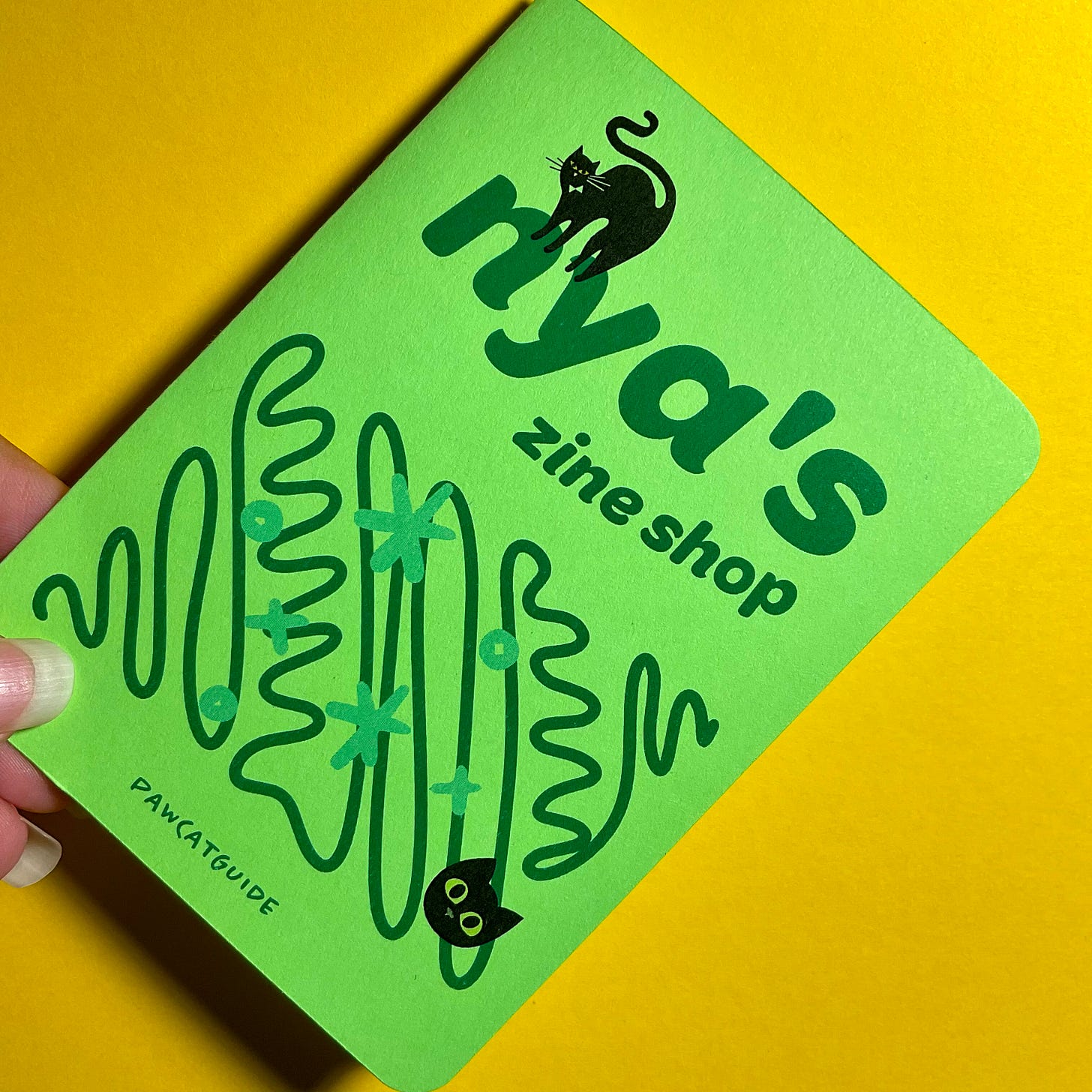 A close-up of a green zine cover with a black cat illustration. The title reads, 'Nya's zine shop' with a smaller text at the bottom reading 'PAWCATGUIDE.' The zine is held against a bright yellow background.