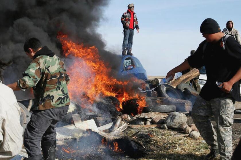 A group of rioters surround a vehicle set on fire, with one standing on top of a vehicle in the background. A group of rioters surround a vehicle set on fire, with one standing on top of a vehicle in the background.