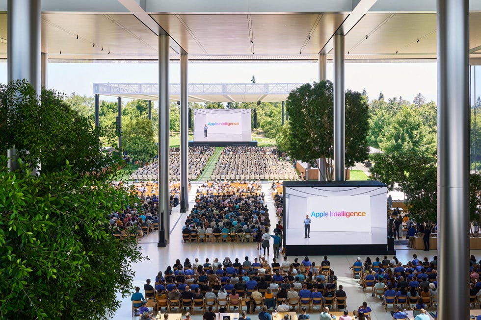 An overhead shot shows developers watching the keynote presentation on large screens in Caffè Macs at Apple Park. 