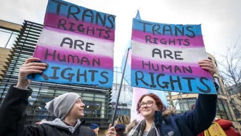 PA Media Two people holdings signs above their heads at a protest, photographed from a low angle. The signs say "trans rights are human rights". The person on the left is wearing a dark jacket and light grey hat. The person on the right has red hair and glasses, and is wearing a blue jacket and grey patterned scarf. People and UK government office buildings can be seen in the background. 