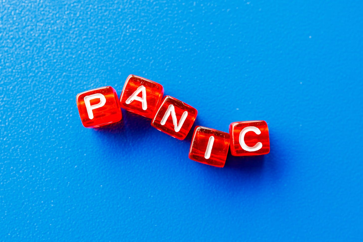 A picture from above of six red dice-like cubes with white letters on them, arranged to spell out the word "panic" in a lopsided but readable way. They are sitting on top of a blue textured background of some sort.