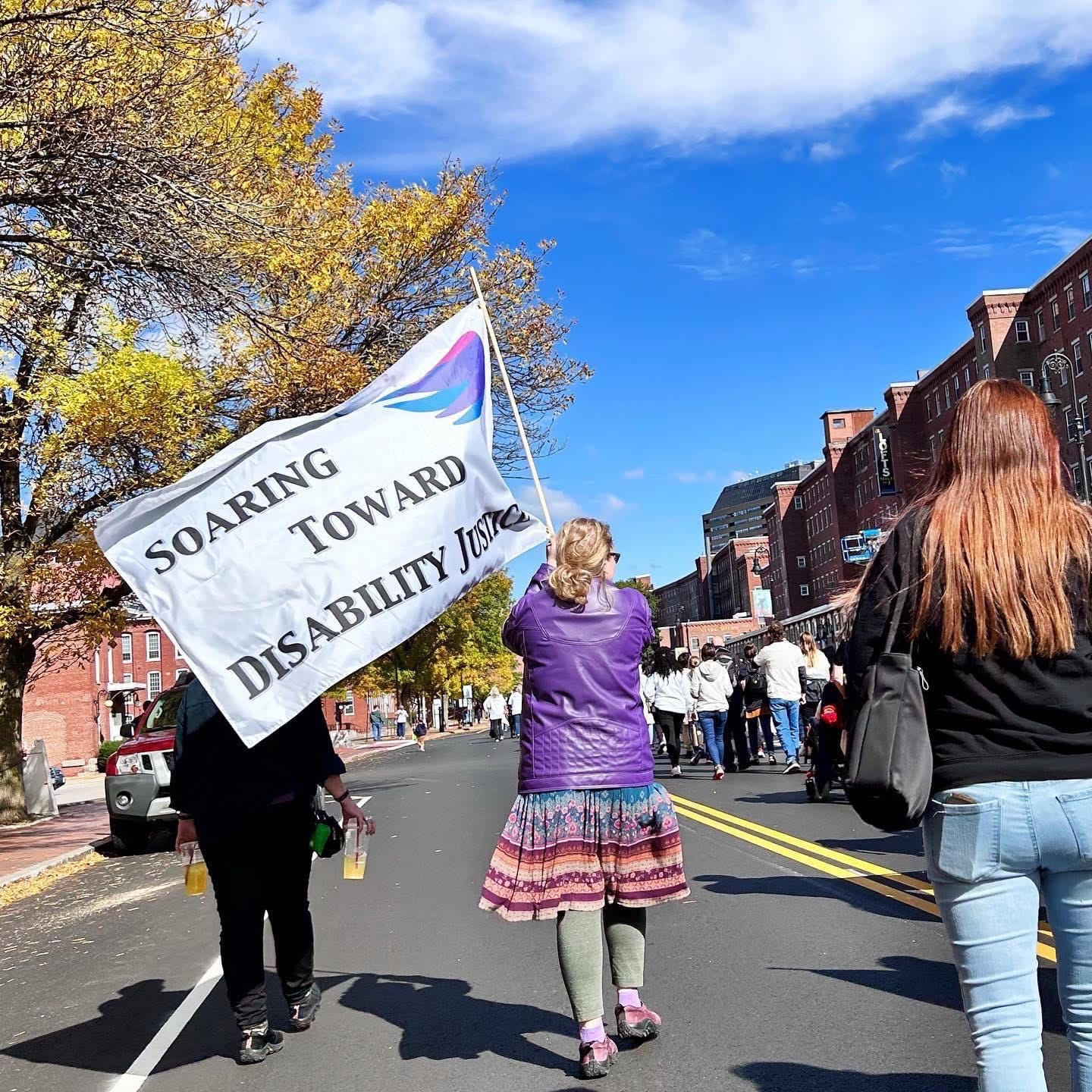 In the middle of a street where a parade is marching, a white woman holds a flag flying behind her that says Soaring Toward Disability Justice.