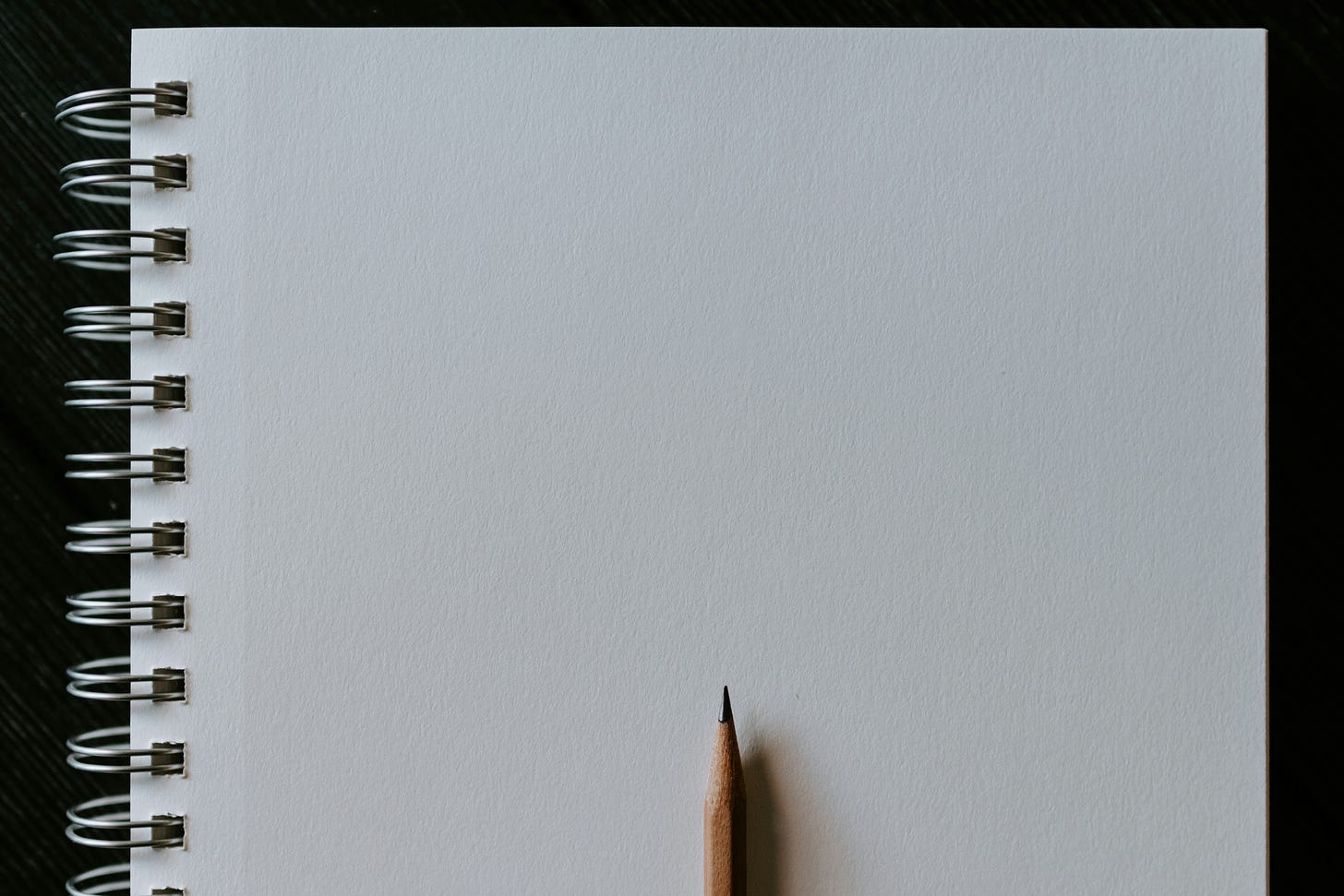 A blank white page of a notebook with spiral binding, and a sharpened pencil placed on top of it.