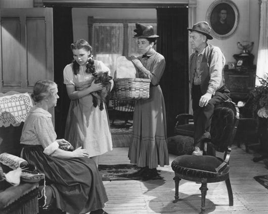 Dorothy está de pé, com seu cachorro, totó, no colo. Ela conversa com uma senhora sentada no sofpa, com mais duas pessoa de pé ao seu lado. A cena se passa em uma sala de estar e é em preto e branco.