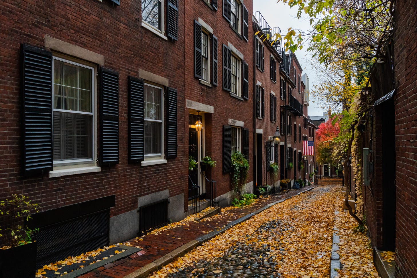 A cobblestone side street in Boston covered in yellow leaves. On each side are brick rowhouses.