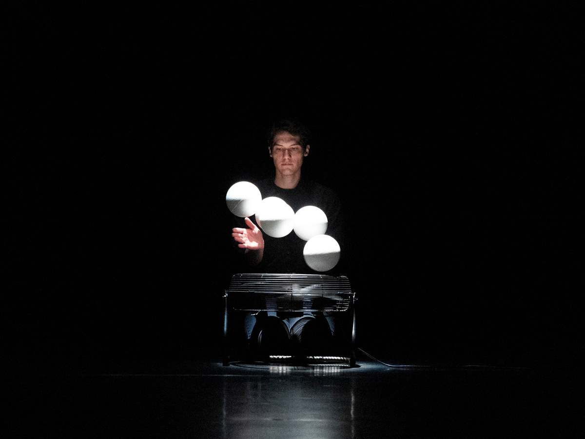 Photograph of a man sitting on a darkened stage behind an electrical fan that blows polystyrene balls in the air