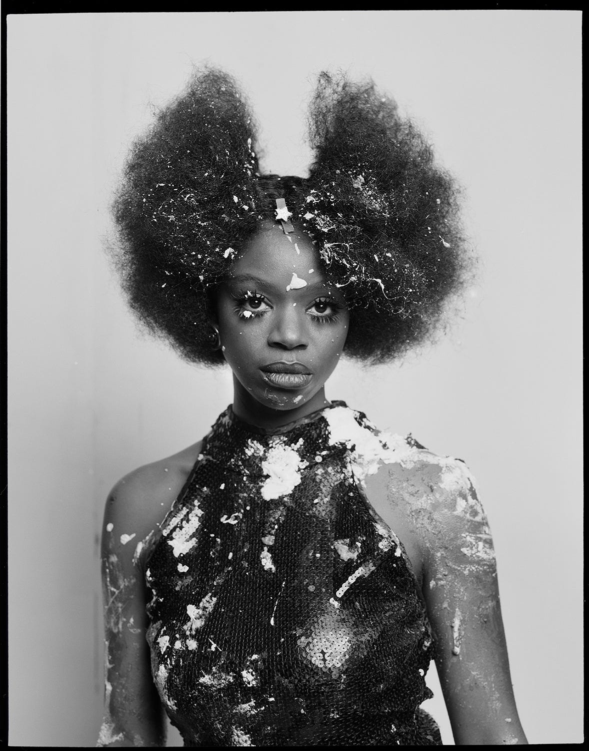 A black and white photographic portrait of a figure staring away from the camera lens. She wears a sequined turtle neck and her hair is in a brushed out afro parted in the middle. There is white paint splattered across her figure. 