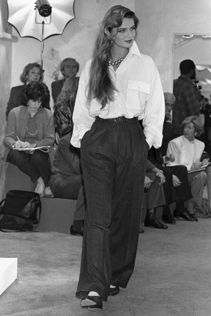 A model wears an oversized whit shirt with a turned up collar and slouchy, high-waisted black pants walks a spectator-lined runway while keeping her hands in her pockets.