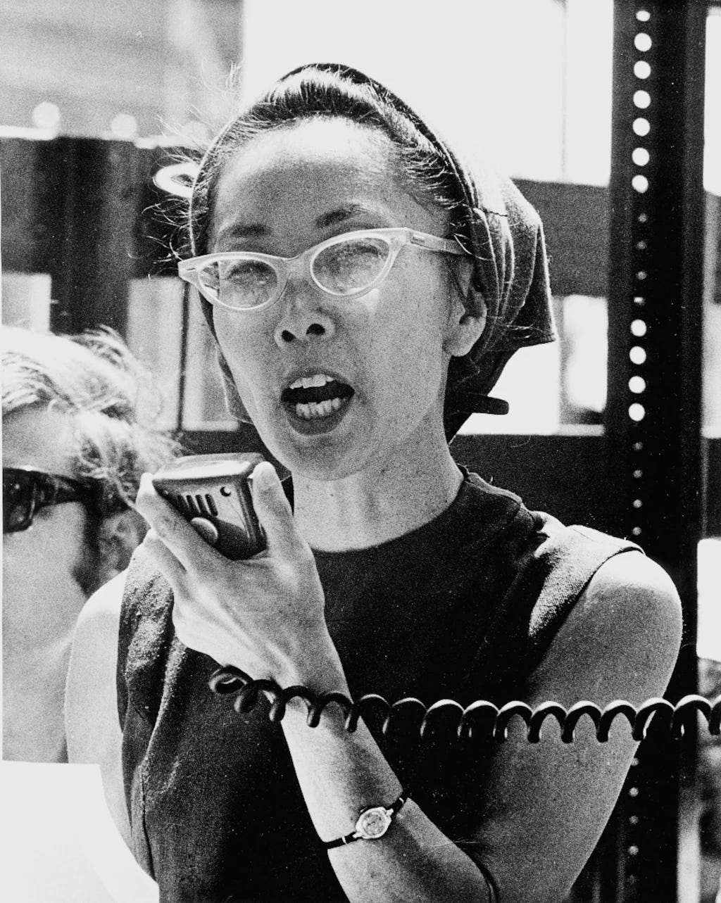 A black and white photo of a Japanese American woman wearing pointy eyeglasses and speaking into a microphone at a protest