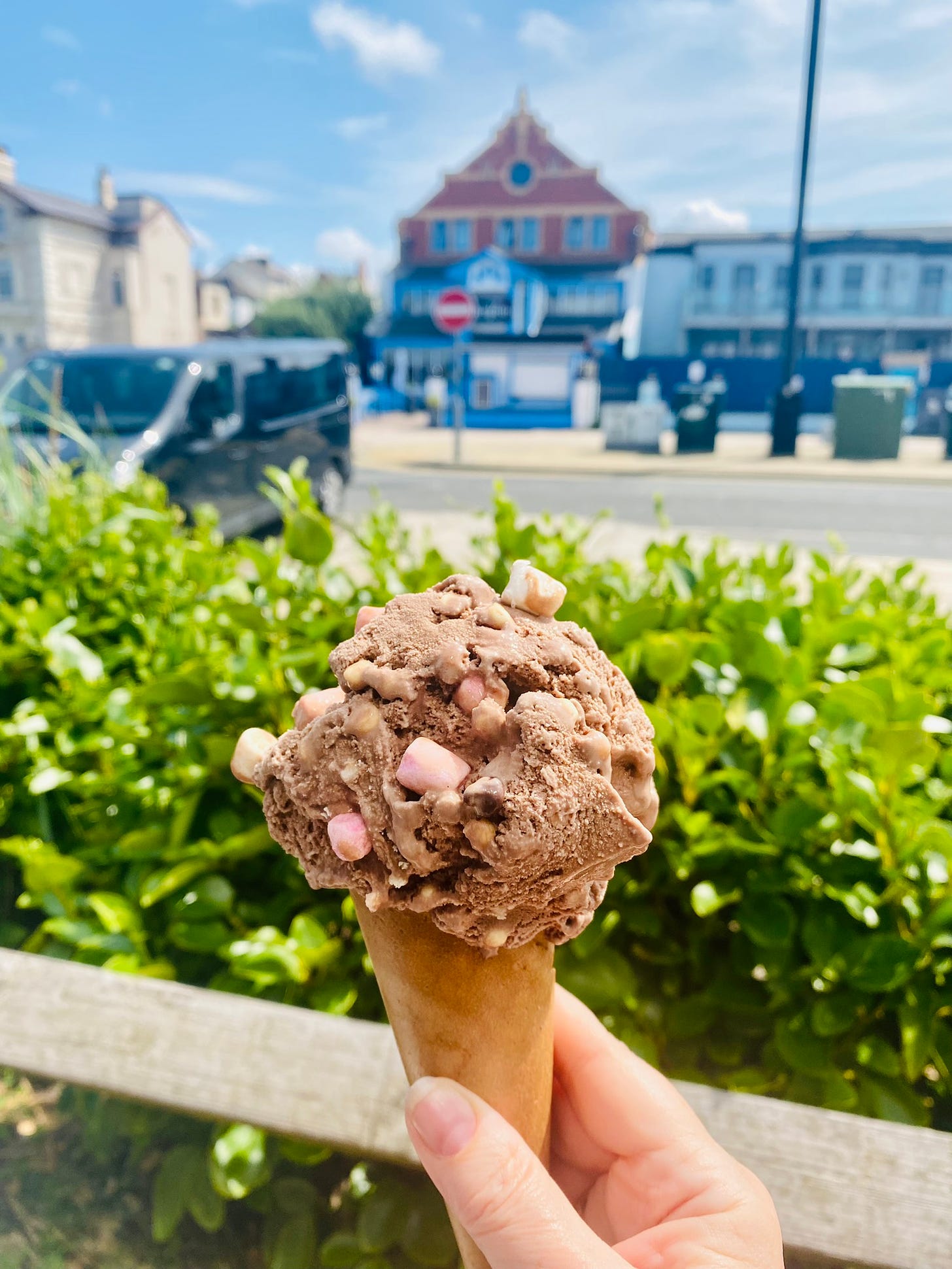 My hand holding a rocky road ice cream in a cone, outside in the sun with a green leafy hedge and a seaside street in the background.