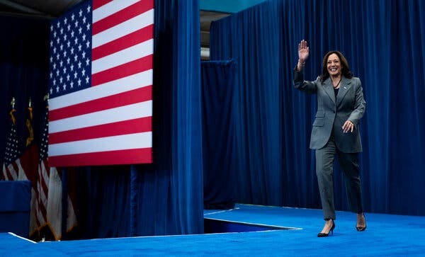 Ms. Harris, wearing a gray suit, waves as she crosses a blue-carpeted stage with an American flag hanging nearby.