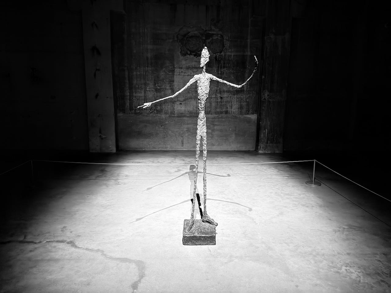 GPT-4o: A black and white photograph of a tall, thin, abstract sculpture of a human figure standing on a small rectangular base. The sculpture is made of a rough, textured material and is displayed in a dimly lit, industrial-style room with concrete floors and walls. The figure has elongated limbs and is posed with one arm extended forward and the other slightly raised. A spotlight casts a dramatic shadow of the sculpture onto the floor behind it. A rope barrier surrounds the artwork. GPT-4o: A black and white photograph of a tall, thin, abstract sculpture of a human figure standing on a small rectangular base. The sculpture is made of a rough, textured material and is displayed in a dimly lit, industrial-style room with concrete floors and walls. The figure has elongated limbs and is posed with one arm extended forward and the other slightly raised. A spotlight casts a dramatic shadow of the sculpture onto the floor behind it. A rope barrier surrounds the artwork.