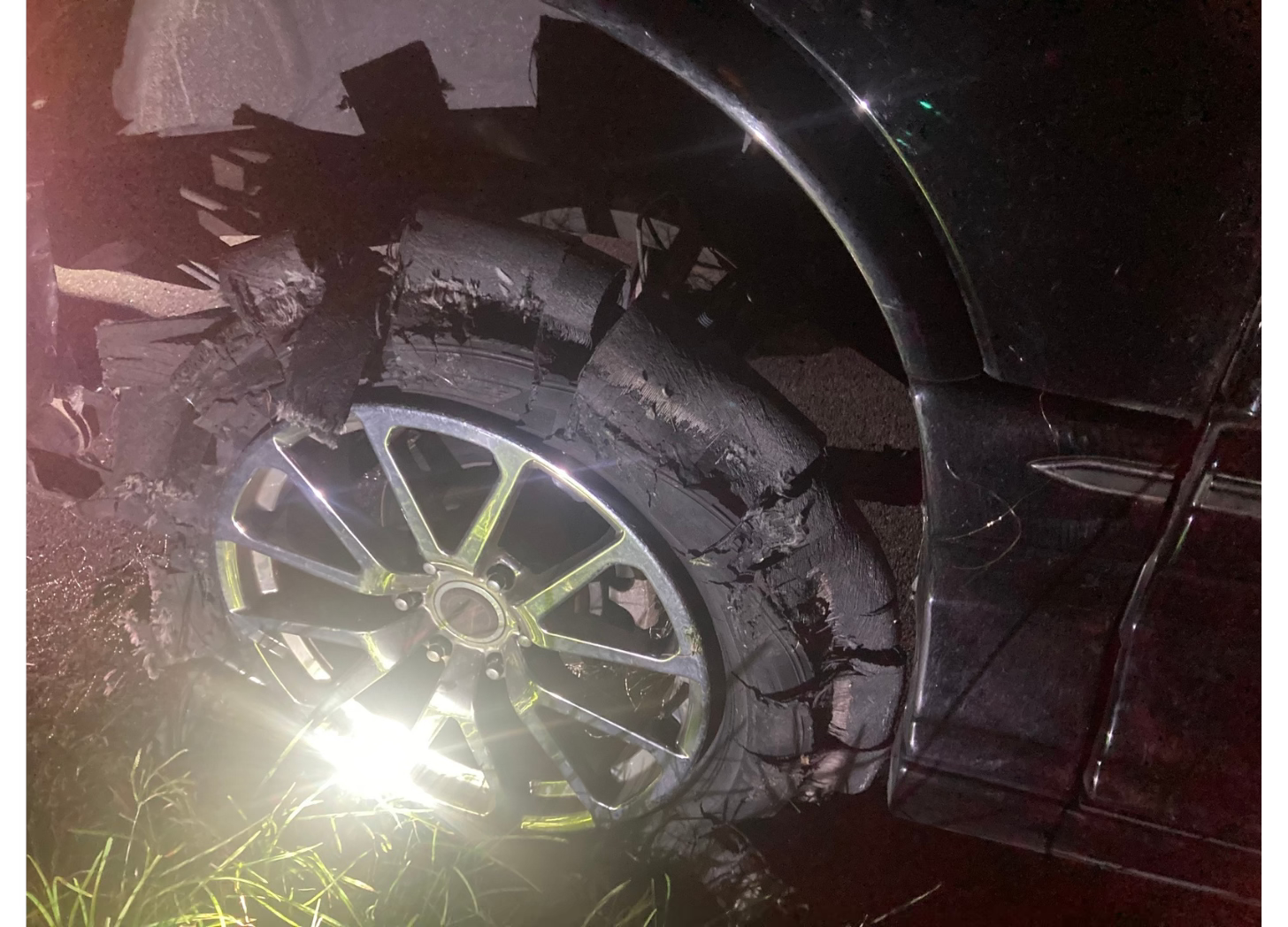 A nightime photo of the close-up of a shredded, flat, very large tire on a black van. A flashlight is shining on it making a glare. 