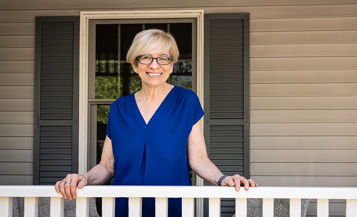 Chai Feldblum standing in a blue short-sleeve blouse, in front of a beige house. Her hands sit on a white fence that is directly in front of her.