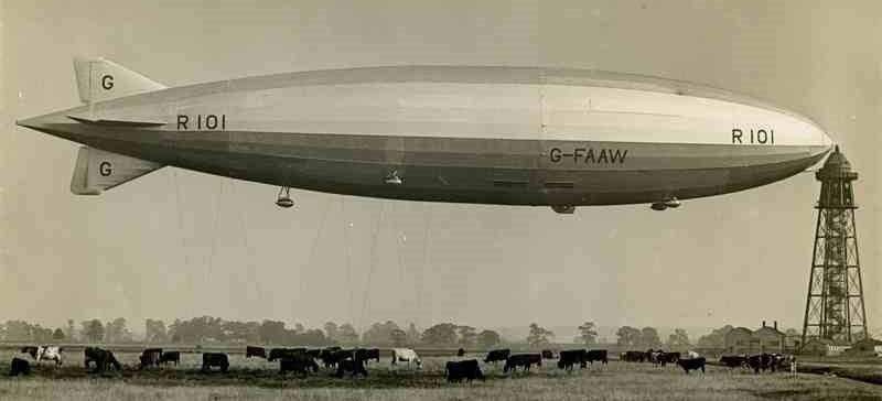 The airship R101 at its mooring mast casting shade on a field of cows.