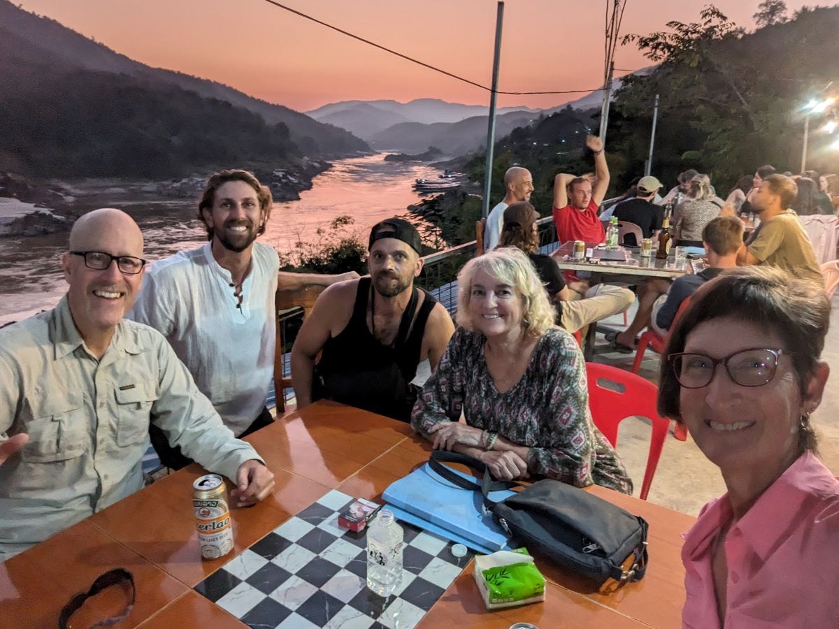 five people sitting at a table overlooking a river, with the sunset making layers of the mountains behind them