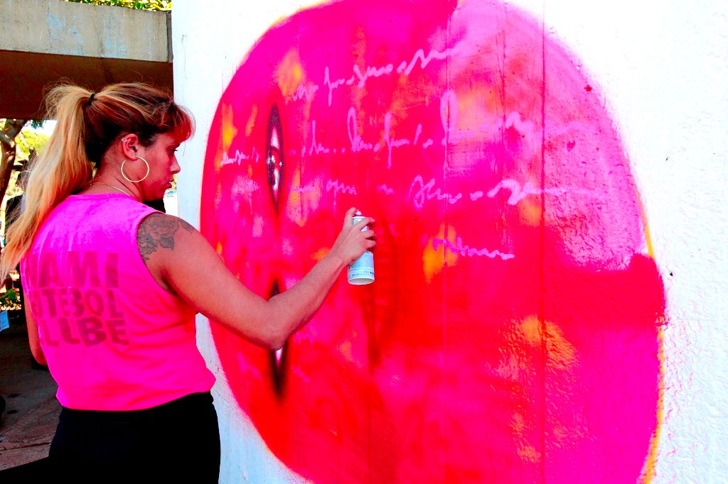 Panmela Castro (Anarkia Boladona) does a live graffiti performance during the Funarte Prize for Women in Visual Arts in Brazil. The artist is poised with a can of spray paint in front of a large painting of a sideways, cartoonish face with white script superimposed over the pink and red circle of the face.