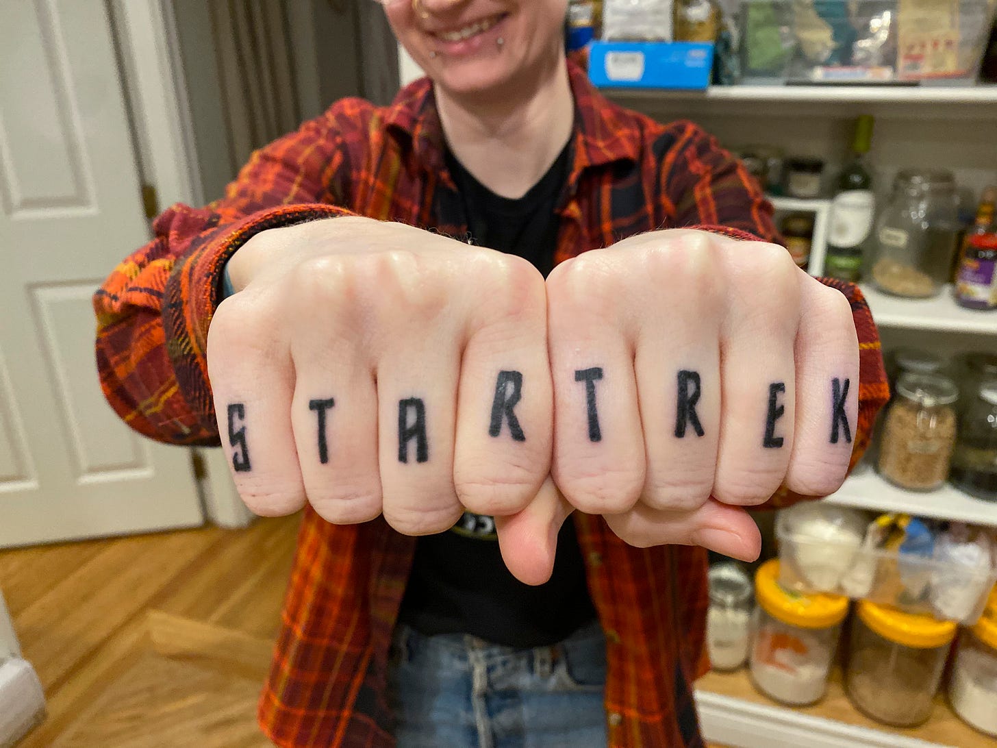 A close-up of my knuckle tattoos, which read 'STAR TREK' in black in the classic font. I am standing in front of our pantry shelves wearing a black t-shirt and an orange plaid shirt overtop. I am smiling just at the top of the frame and the rest of my face is cut off.