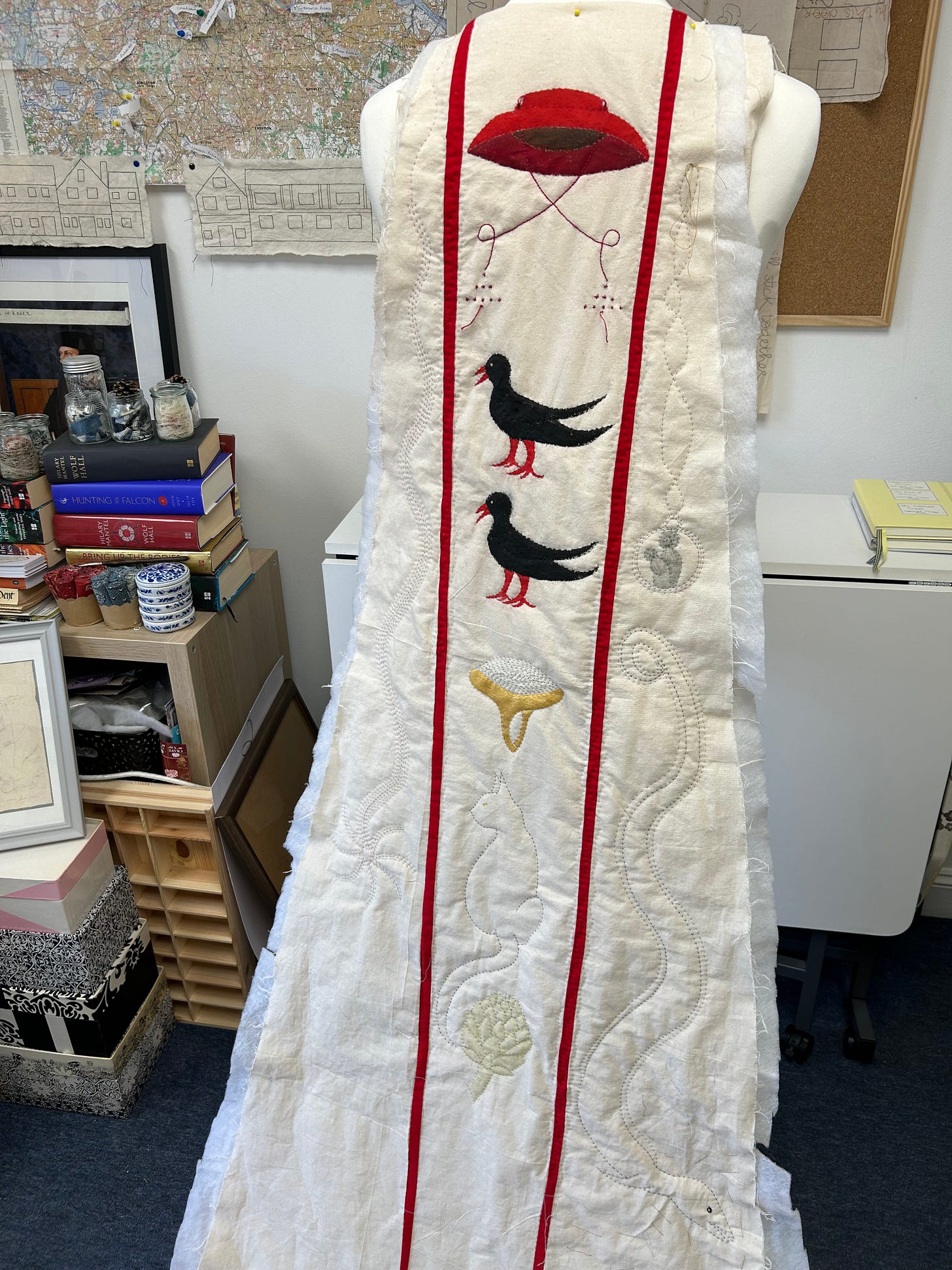 A partially completed long cloak back hanging off a dressmakers’ form. The cloak is cream coloured with various motifs stitched on - a cardinal’s hat, two black birds, an artichoke, a comet, an eel, a cat, a holy medal, a turquoise ring. There are red strips dividing three sections.