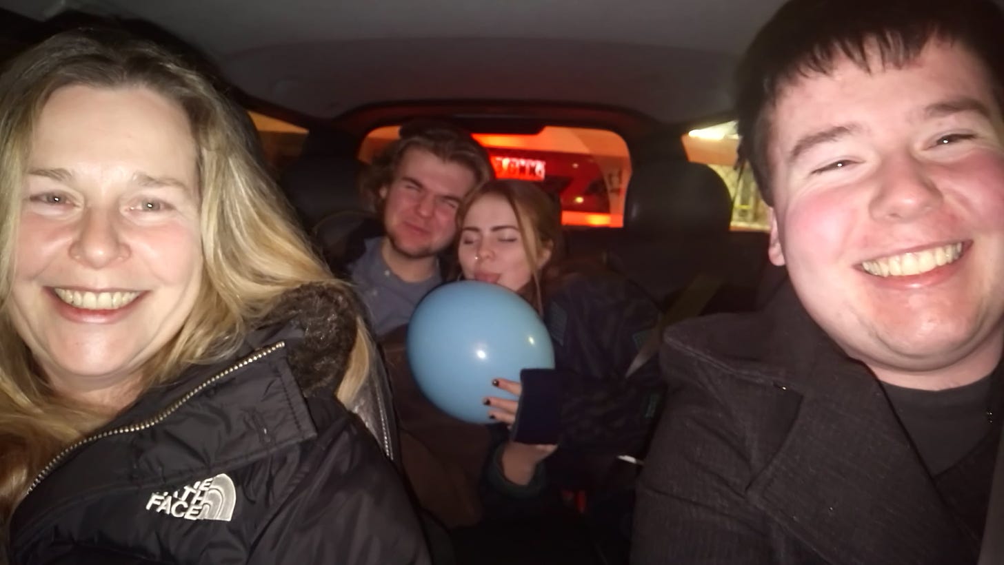 Mother and one son in the front seat of a car at night, another son and female friend with a blue balloon in the back seat--all are smiling widely