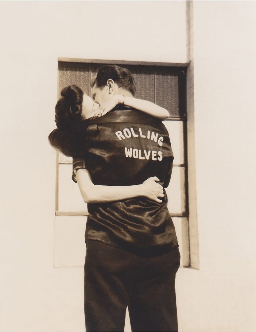A sepia-toned photo likely from the 1950s. A greaser couple kisses. The man's back is to the photographer and his black jacket reads "Rolling Wolves" on the back.