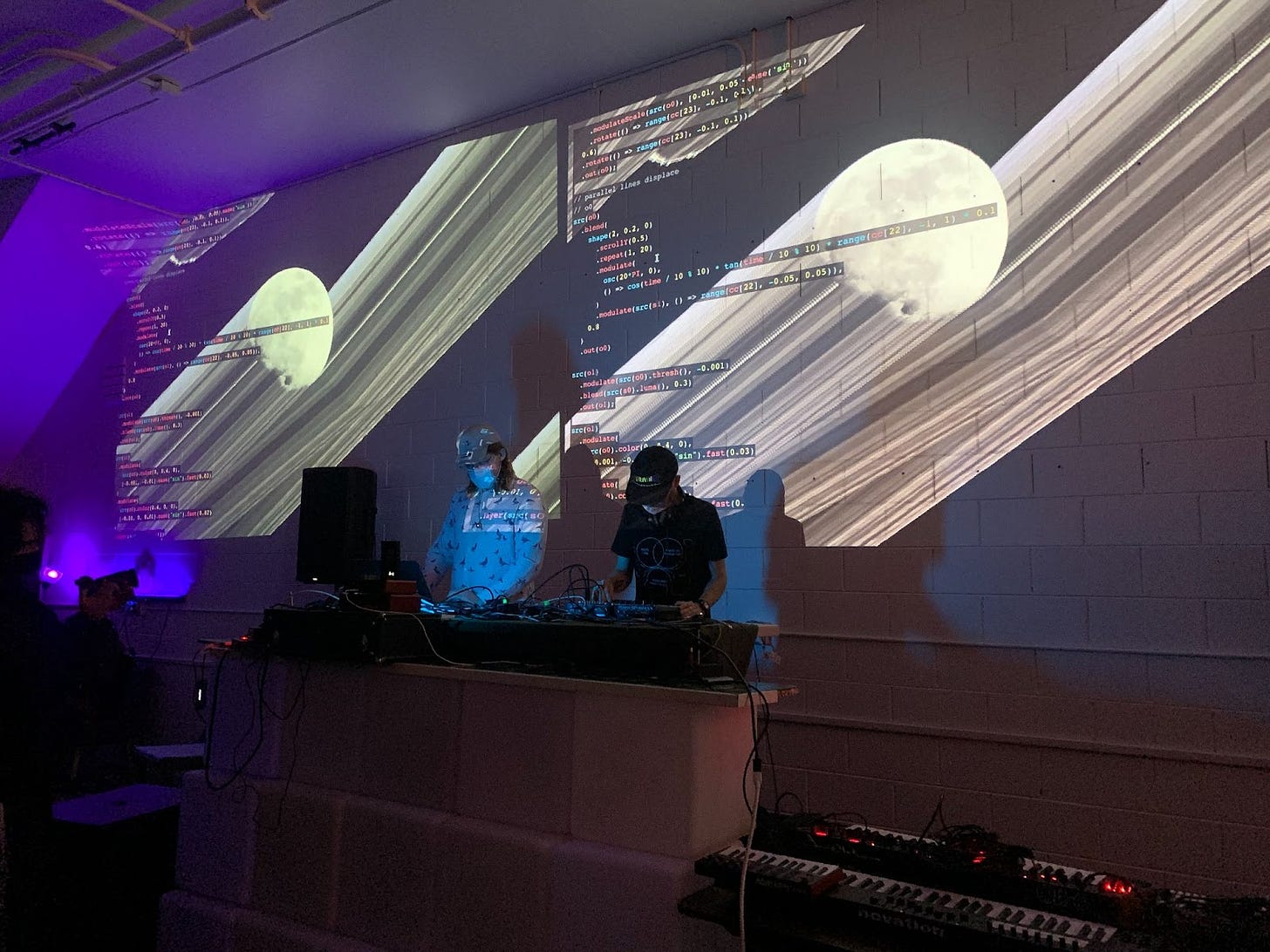 Two DJs mix in the front of a dark room, with 2 screens of code projected on the wall behind them