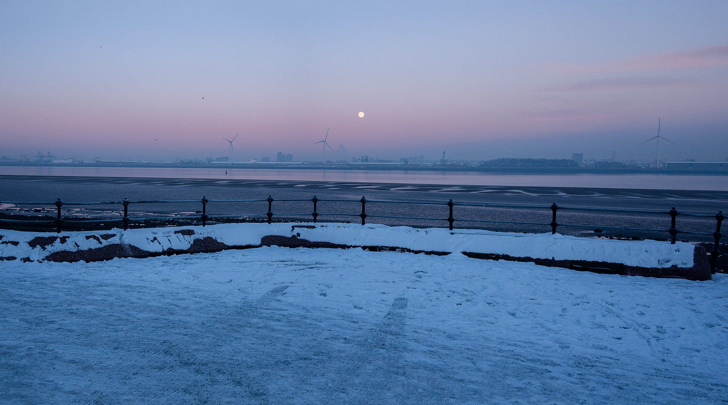 Just after sunset, at dusk, there is a pink glow in the sky. The moon is rising and there is snow covering the promenade by the River Mersey.