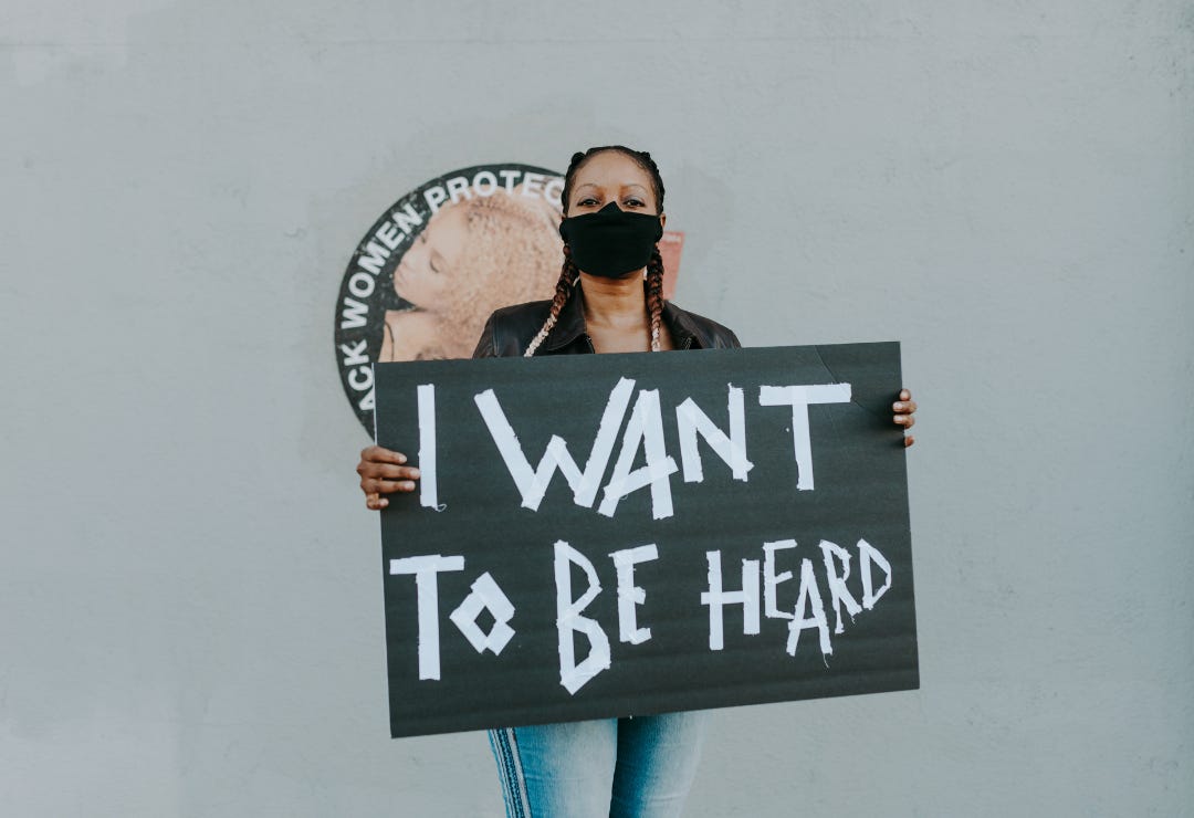 Black woman wears a mask covering her mouth while holding a placard that says, "I Want to be heard"