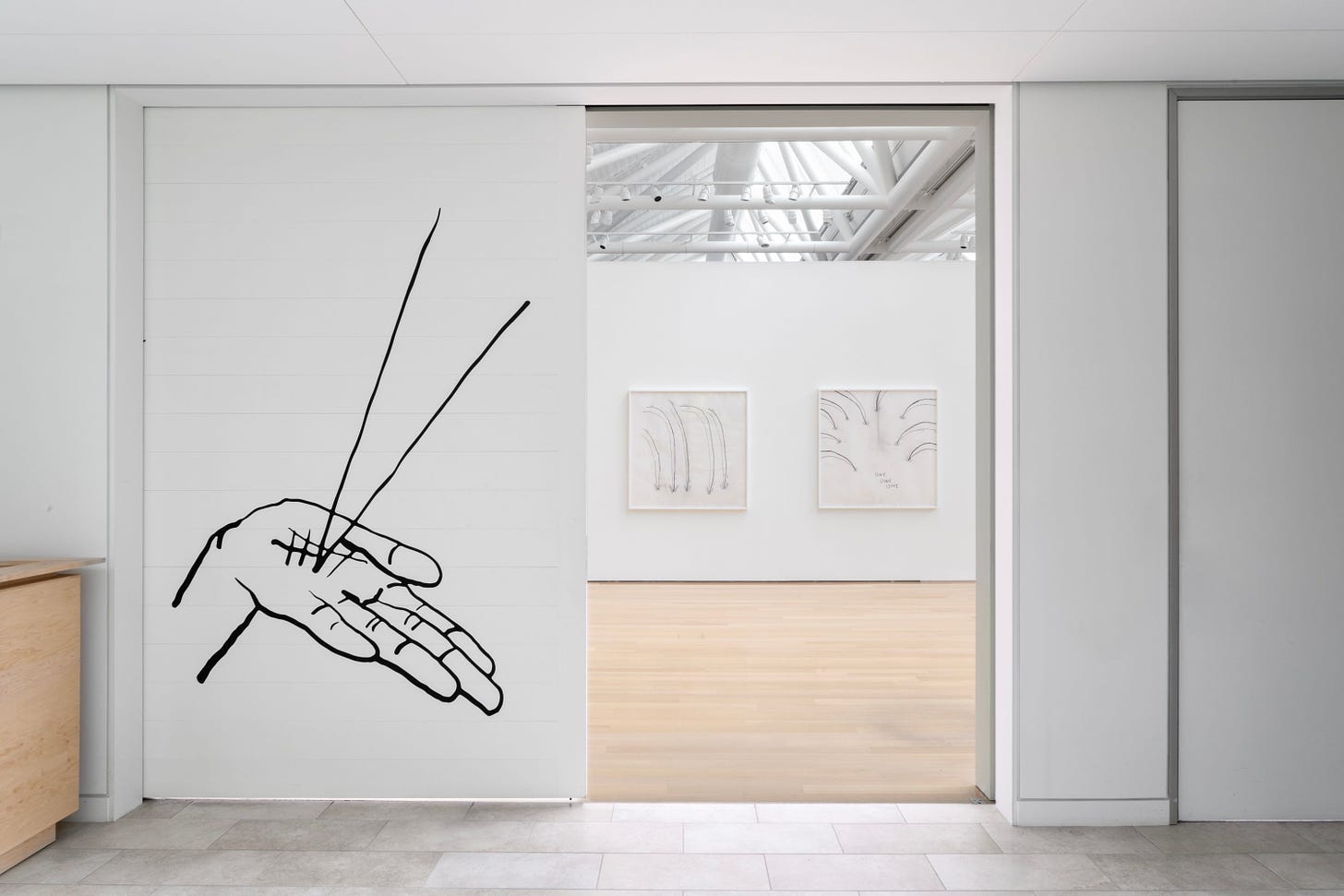 Installation view of a doorway inside a white-walled gallery. On one door, there’s a drawing of a hand with two lines leaping out. Inside, there are two works on a wall, too far away to know detail.