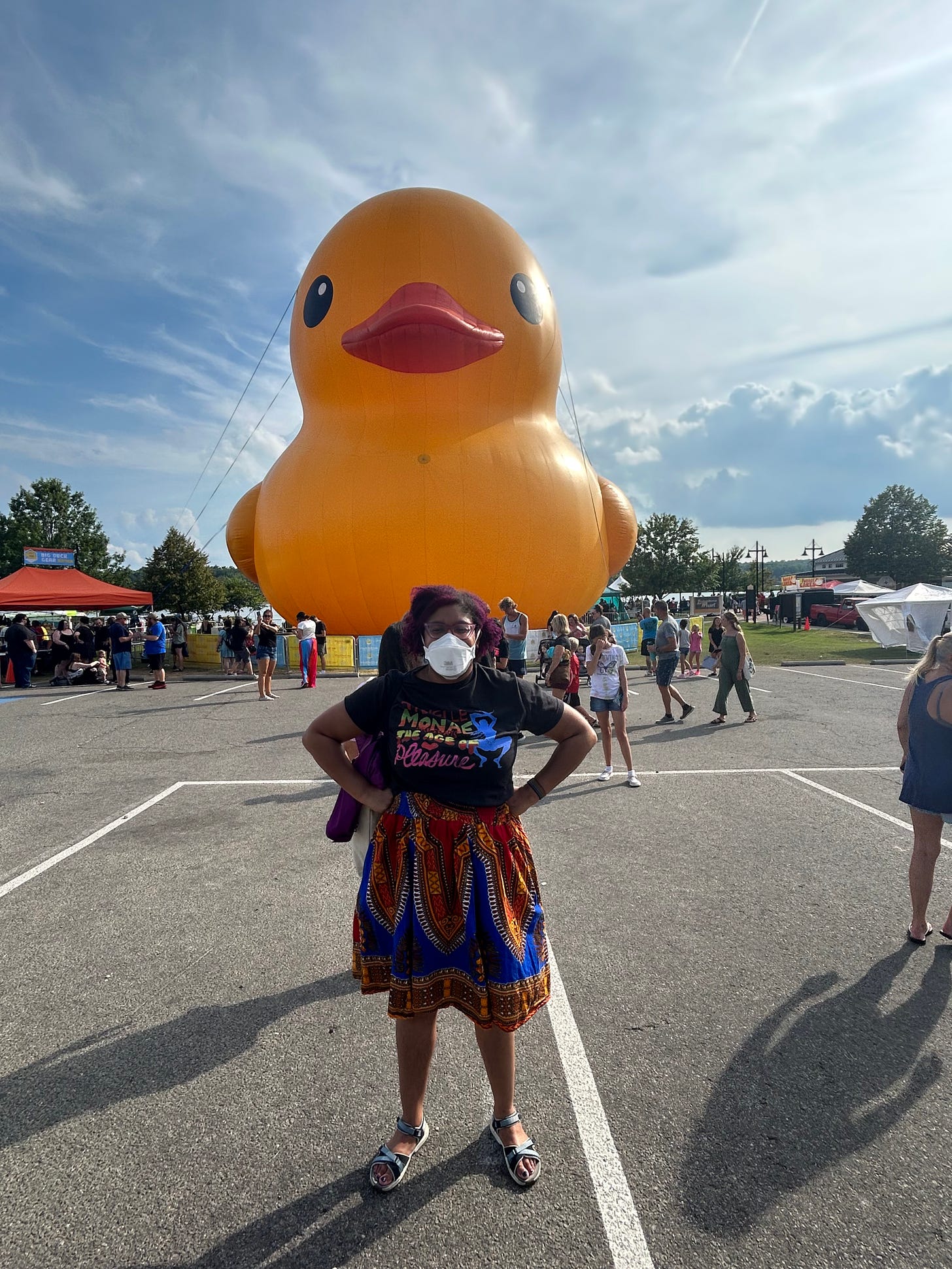 Kristen is standing, masked and arms on hips, in a Janelle Monae t-shirt,in front of a large inflatable object in the shape of a yellow plastic duck