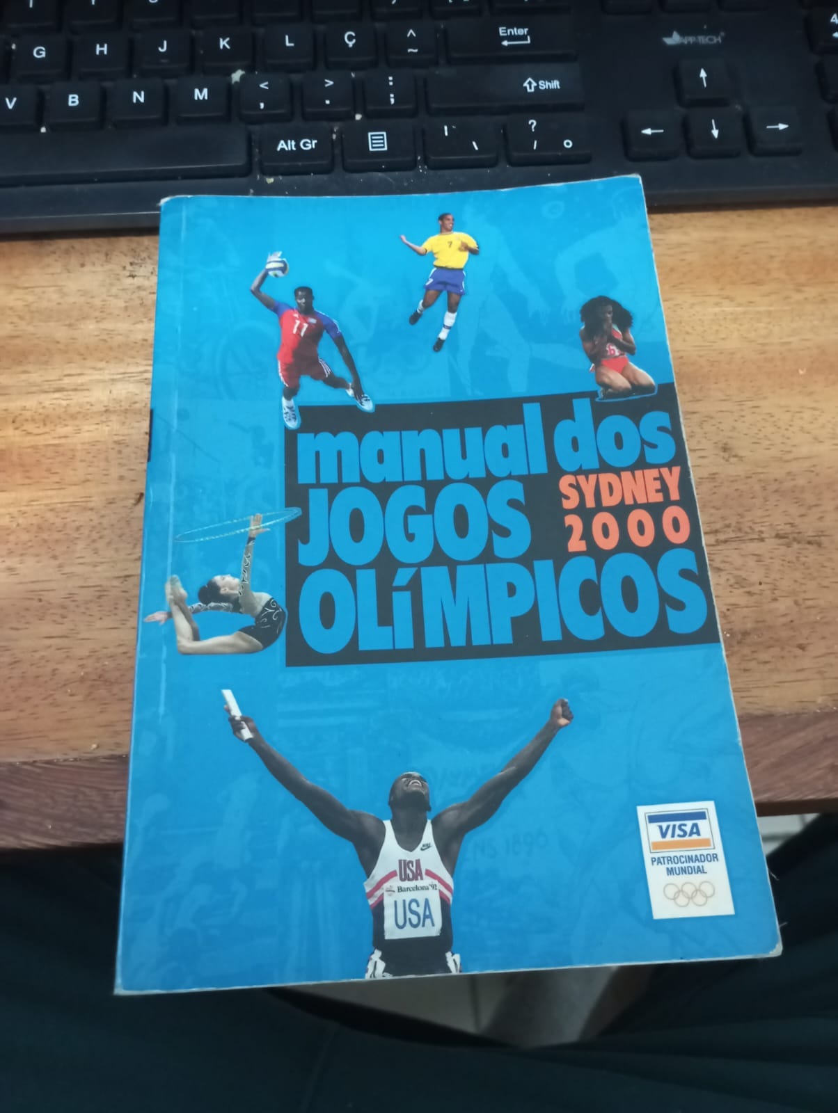 Imagem do Manual Visa dos JogosOlímpicos sobre Sydney-2000. Um libro de capa azul, com dizeres da capa e imagens de altetas espalhadas