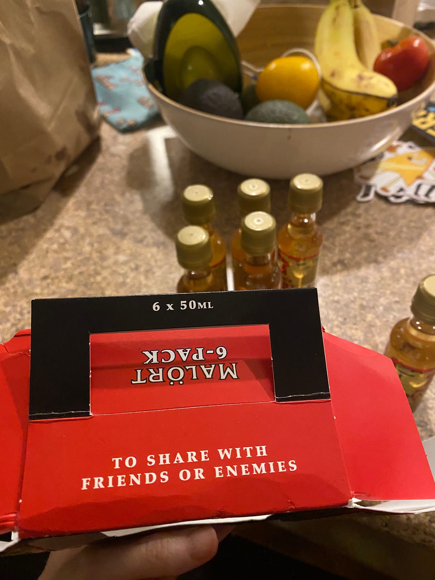In the foreground, a red paper box with a black handle that says "6x50mL". One side of the box reads 'to share with friends or enemies" in white block letters, and the other side reads "6-pack Malört". Behind the box are the 6 little bottles, still full, with gold lids.