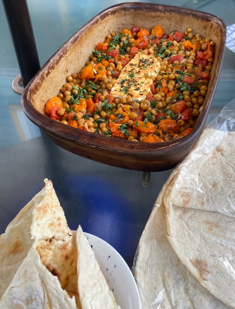 A stoneware dish of the chickpea & tomato baked feta described above, with chili flakes and chopped herbs all over the top. In the foreground is a package of taftoon, and a torn piece of it resting on a side plate.