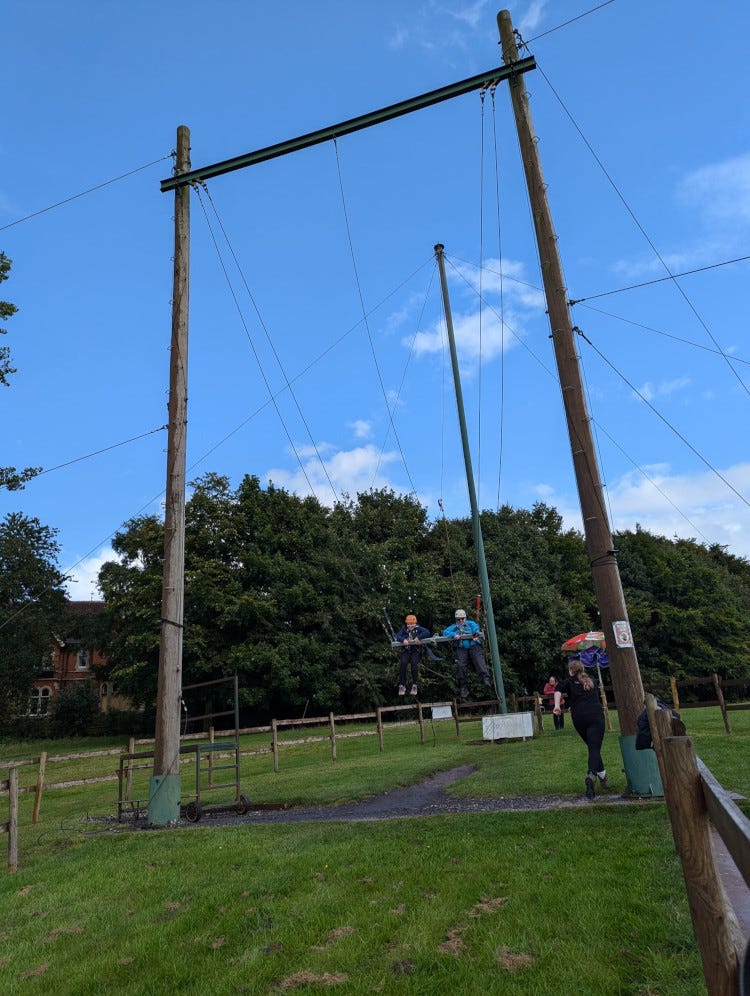 Being winched up the giant swing, wearing my big blue waterproof because it was raining when we walked down, although the sky is now bright blue and sunny. The giant swing is a giant wooden frame covered in wires; four of them go down to a metal bar which Maddi and I are harnessed to and another wire, leading to a pole behind us, is hauling us up towards the sky.