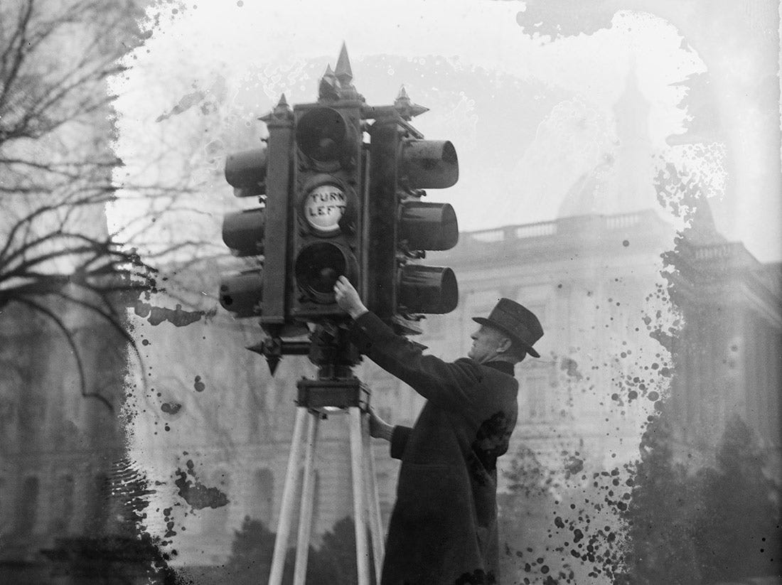Black and white photo of a man in a hat and coat adjusting an old-fashioned set of traffic lights (1926)