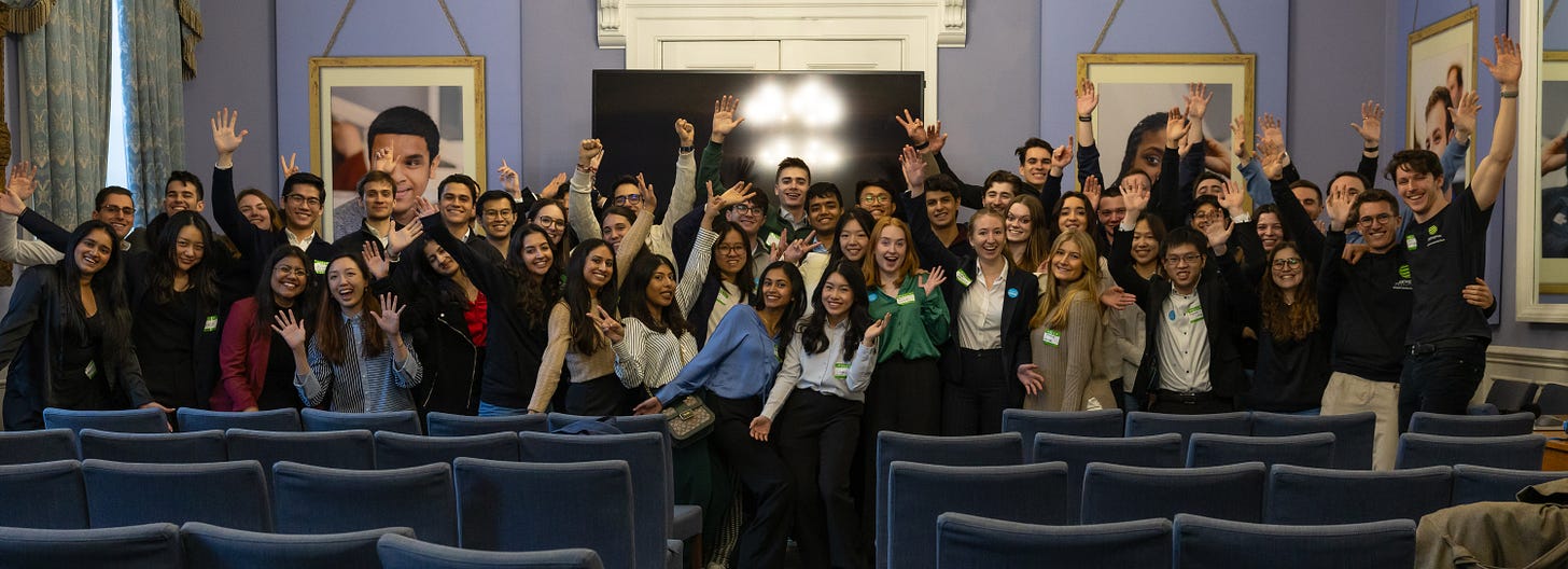 A large group of students standing behind a large screen television, in front of a seat of blue chairs, waving their hands in the air and smiling.