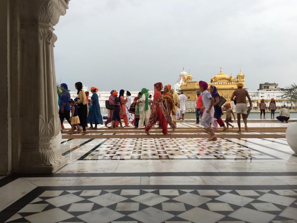 View of a marble floor and pillar in the foreground, on the grounds of the Golden Temple. People wearing Indian and western attire walk up ahead, with the golden inner sanctum visible in the distance. The sky is overcast.