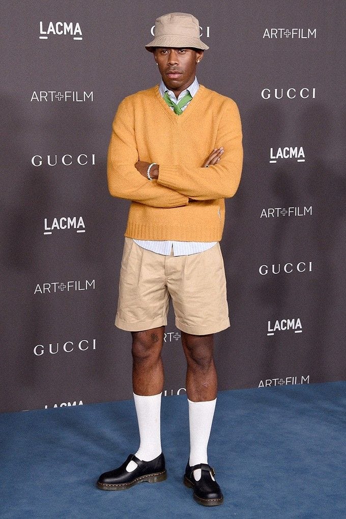 This may contain: a man with his arms crossed standing in front of a blue carpet and wearing a yellow sweater